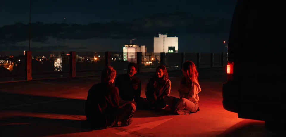 Four people on a rooftop car park at night, lit in red.